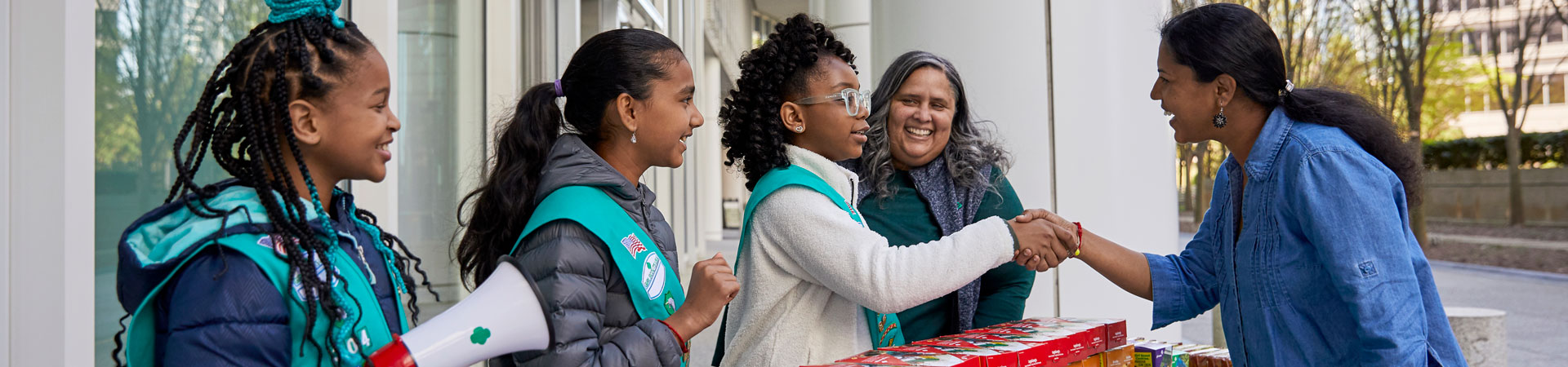  A young adult in a jean shirt shakes the hand of a Girl Scout behind a table stacked with Girl Scout cookies. 